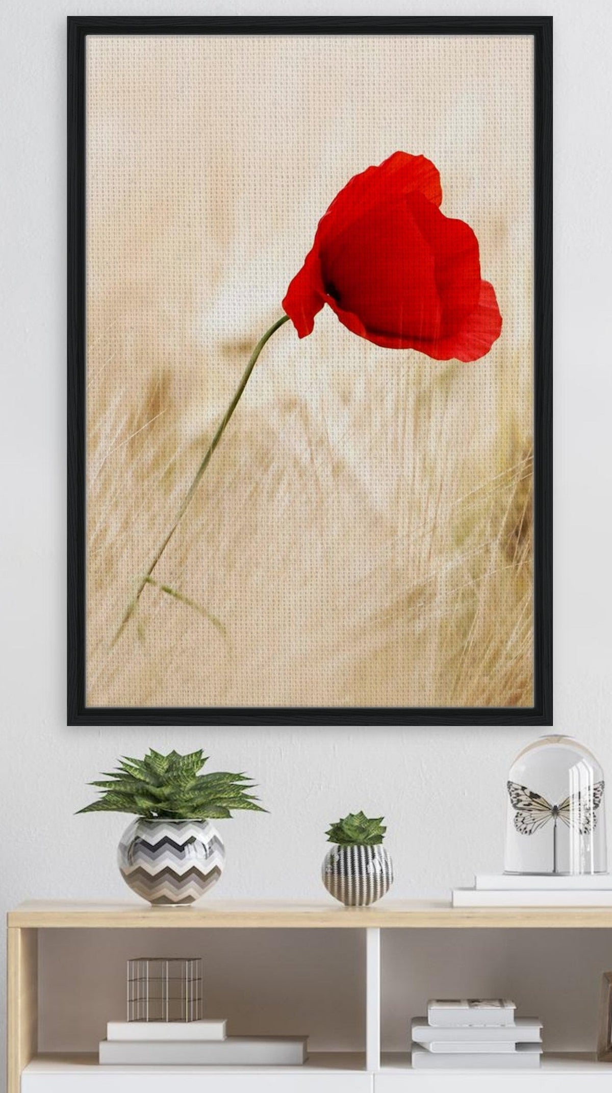 A vertical framed wall art print featuring a single red poppy flower on a textured beige background, displayed above a white sideboard in a modern room setting with a floor lamp and decorative objects.
